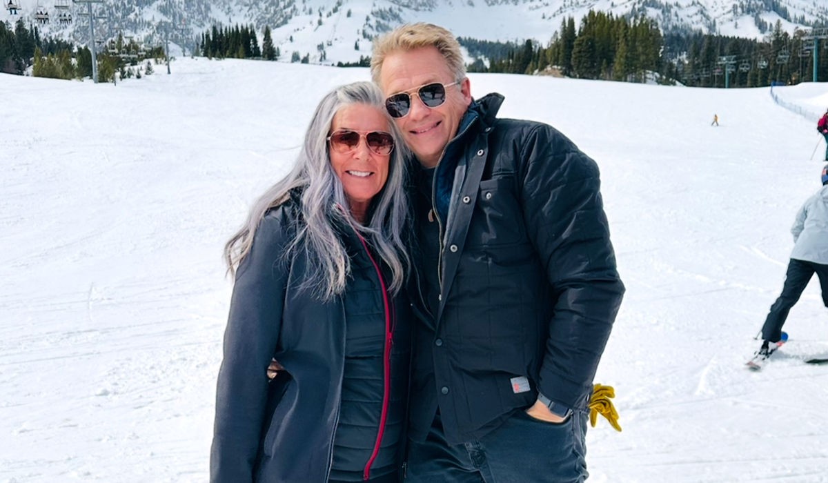Paul and Kelli are wearing winter clothing stand on a snowy ski slope with trees and mountains behind them.