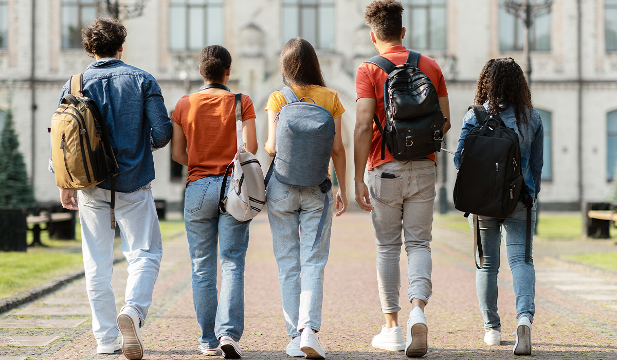 Five people wearing backpacks walk together on an outdoor path toward a large building.