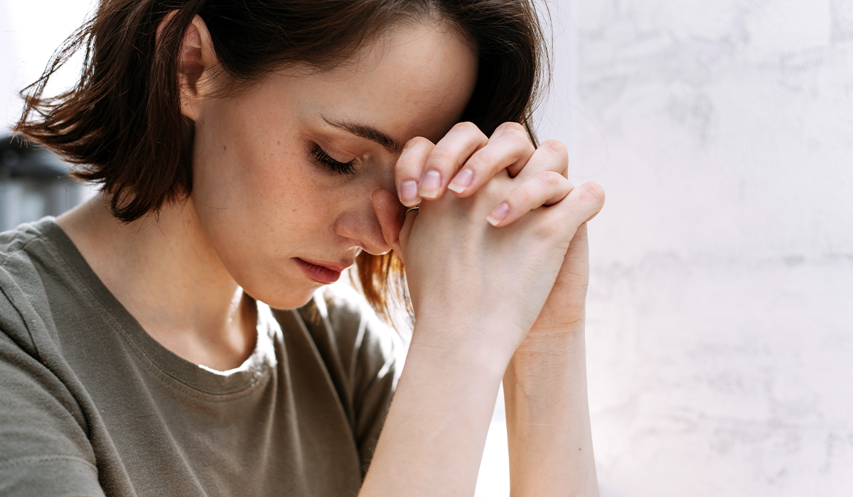 Person sits near a window with head bowed and hands clasped together in front of the face, lit by natural light.
