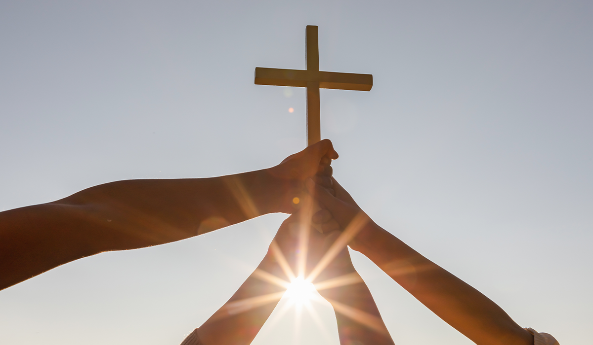 Multiple hands hold a wooden cross raised toward the sky with sunlight visible behind it.