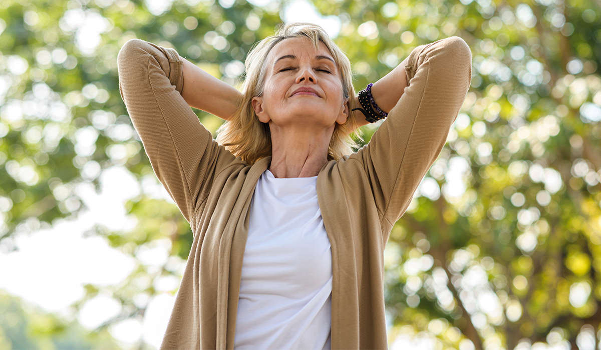 An older woman standing in a forest, taking a deep breath with her hands resting on her head, surrounded by lush greenery and a serene natural backdrop.